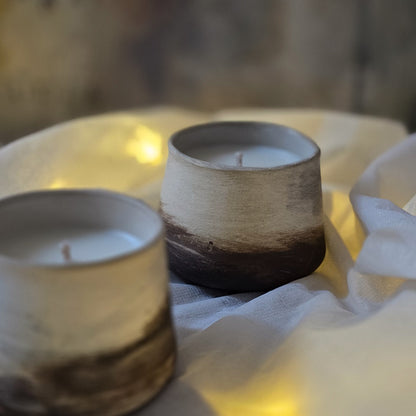 Two small ceramic candle holders with a rustic hand-painted finish, one in warm off-white and brown and the other in beige and dark brown, filled with white wax and set on draped white fabric with warm yellow lighting.