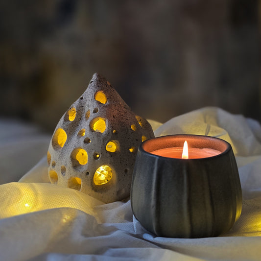 A deep green ceramic candle holder with a lit fragrant, soy candle next to a decorative perforated ceramic seed pod glowing with warm yellow light, set on draped white fabric