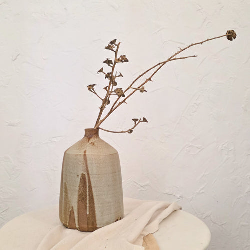 A handmade ceramic vase with a textured, speckled beige and brown glaze, holding a dried brown flower stem, set on a draped cream fabric surface against a white plaster wall.