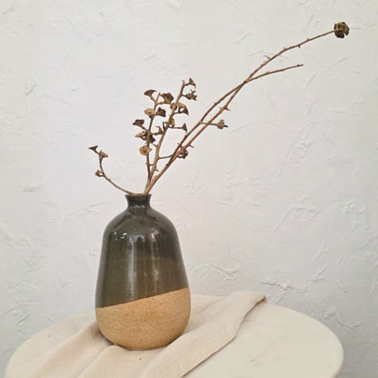 A small ceramic vase with a glossy dark gray upper body and a matte beige lower section, holding dried brown branches, set on a light fabric-covered round table against a textured off-white wall.