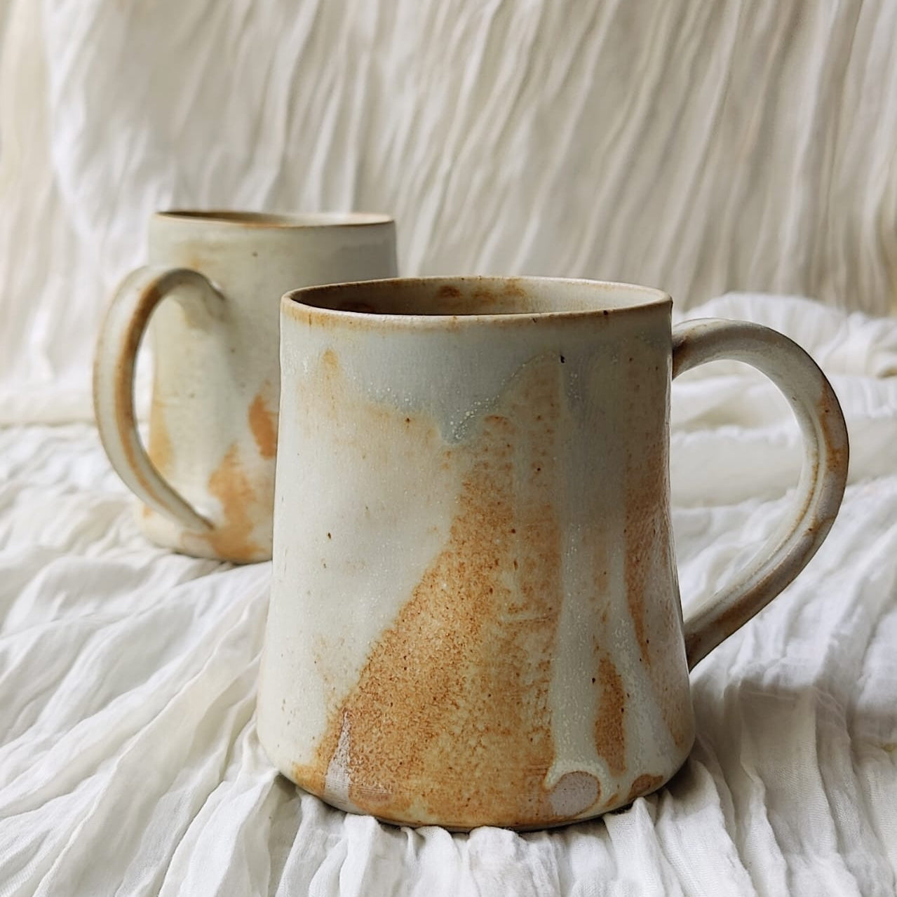 Two handmade ceramic mugs with organic brown and beige glaze variations, displayed on a draped white fabric background.