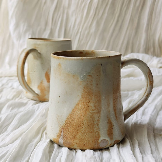 Two handmade ceramic mugs with organic brown and beige glaze variations, displayed on a draped white fabric background.