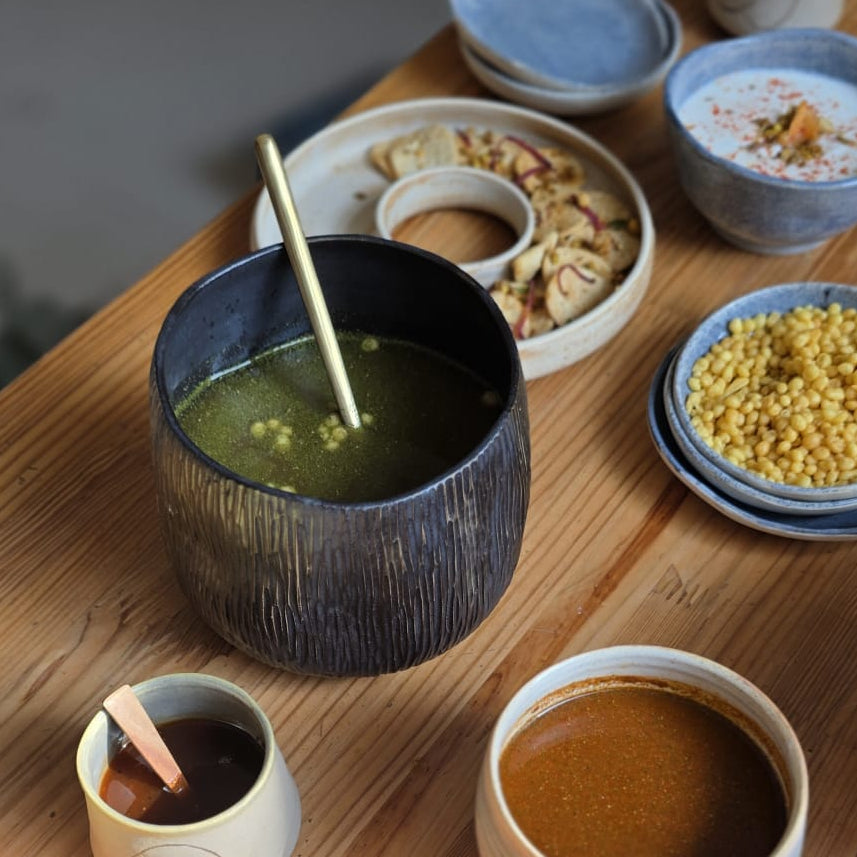 A styled overhead view of a wooden table laden with various Indian snacks and drinks, including a bowl of green chicory leaf soup, a cup of brown chutney, a plate of golden puri, and other small dishes.