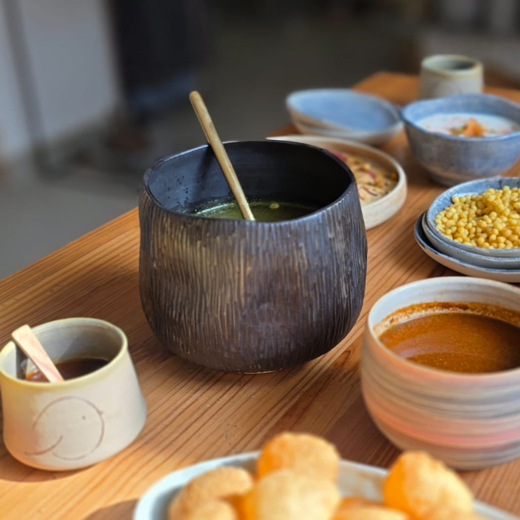 A wooden table set with a large plate of golden fried snacks, a dark textured bowl with a spoon, and several small ceramic dipping bowls and cups in warm natural light.