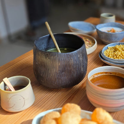 A wooden table set with a large plate of golden fried snacks, a dark textured bowl with a spoon, and several small ceramic dipping bowls and cups in warm natural light.