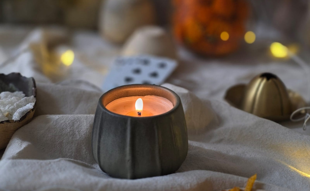 A lit candle in a dark gray ribbed ceramic holder sits on draped cream fabric with warm fairy lights and diwali festive decor softly blurred in the background.