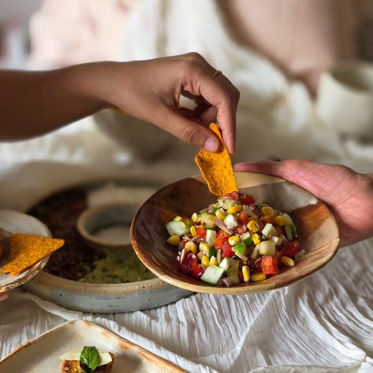 Person dipping a tortilla chip into a bowl of colorful salad with other food items on a table.