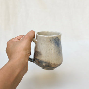 A hand holding a handmade ceramic mug with a rustic, earthy glaze against a soft, light beige fabric background.