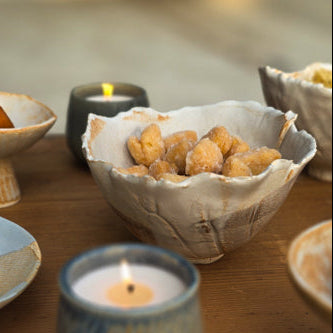 A rustic ceramic bowl filled with golden-brown fried snacks sits on a wooden table next to a lit blue-gray candle, with other bowls and candles blurred in the background.